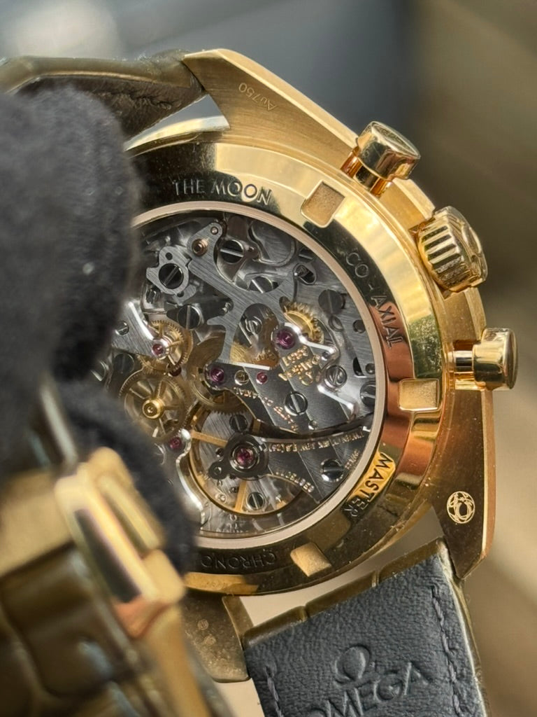 Close-up of a gold wristwatch with a transparent back, featuring the intricate inner mechanics of the Omega 3861 manual winding calibre and engravings around the edge, including "THE MOON," "GOLD," and "OMEGA." Partial view of a gloved hand showcases the 42mm Moonshine™ gold case. The product is an Omega Speedmaster Professional "Moonshine" 310.63.42.50.10.001 by Omega.

