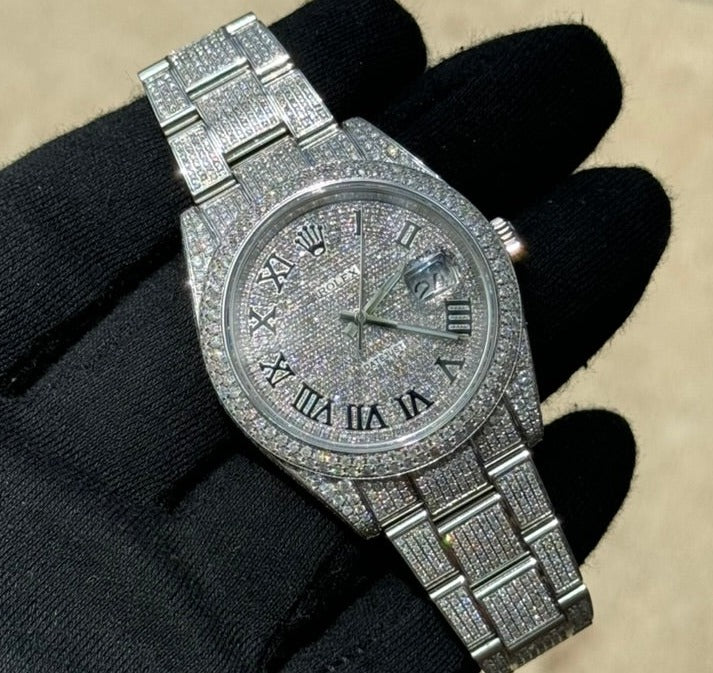 A gloved hand holds a luxury watch, a silver Rolex Datejust 126300 "Bust Down" encrusted with diamonds, featuring Roman numeral hour markers and a date window.