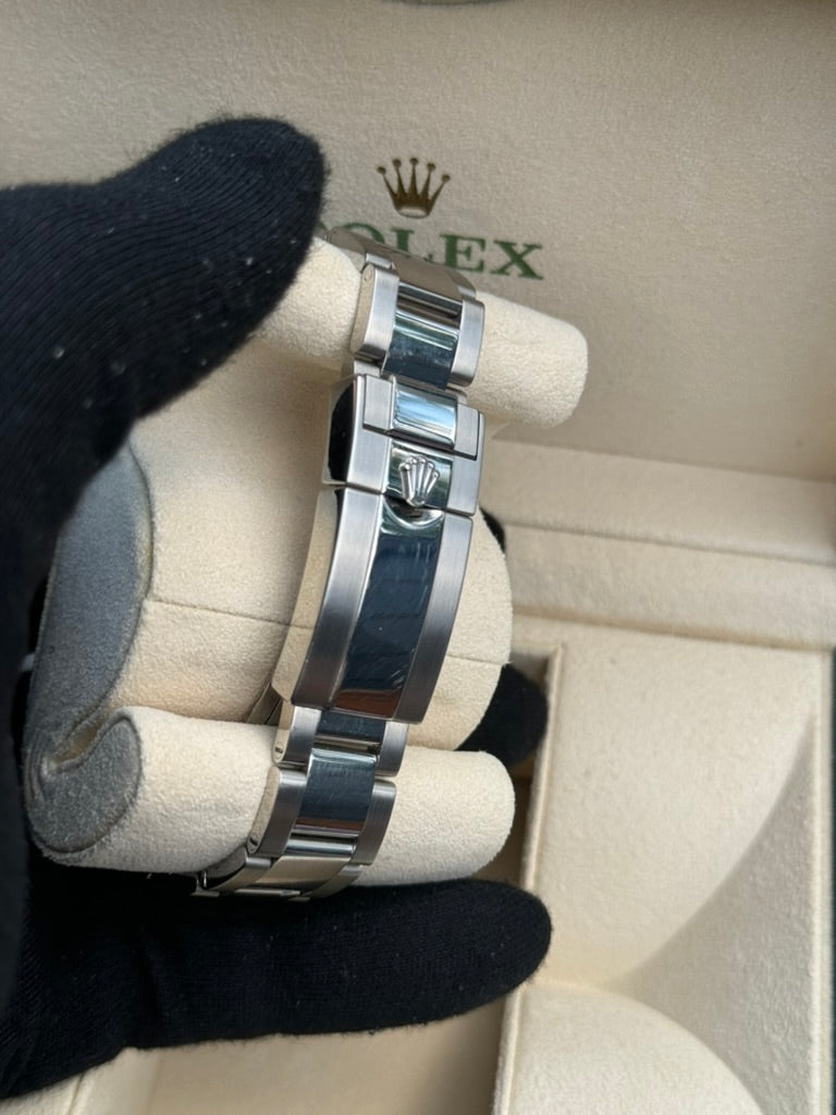 A gloved hand holds a Rolex GMT-Master II 116710LN with a metal band and black ceramic bezel, displayed inside an open beige and green Rolex box.