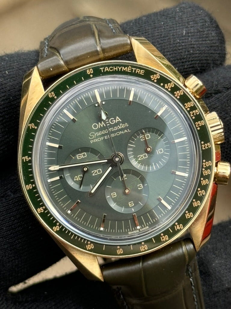 Close-up of an Omega Speedmaster Professional "Moonshine" 310.63.42.50.10.001 wristwatch with a green dial, gold bezel, and black leather strap, featuring the 3861 manual winding calibre, being held by a hand in a black glove.