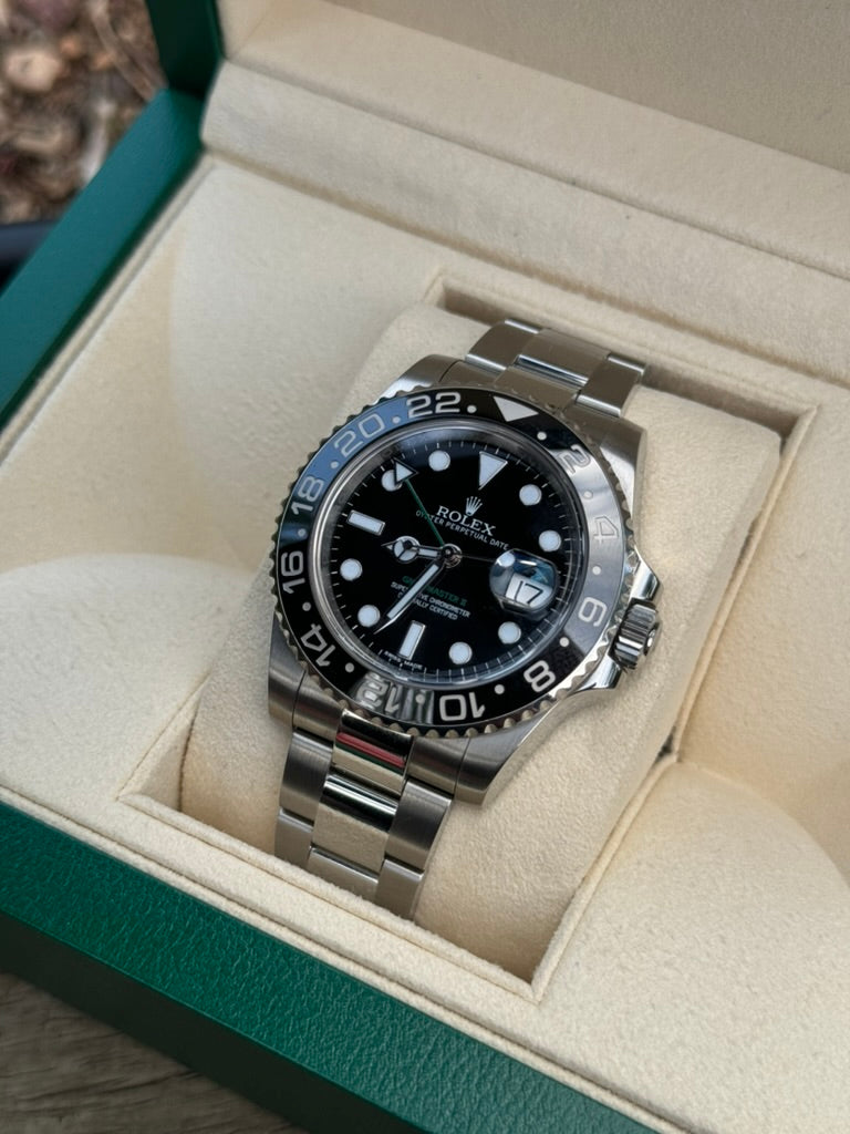 A Rolex GMT-Master II 116710LN with a black ceramic bezel, black dial, and silver bracelet displayed in a green and beige box.