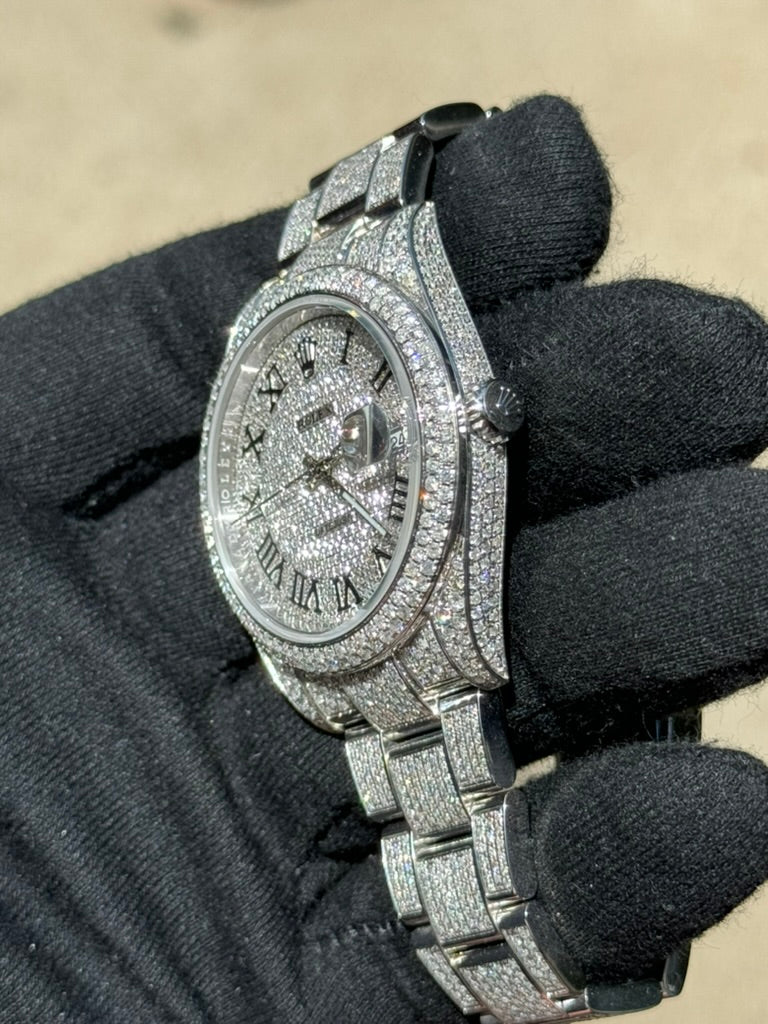 A hand wearing a black glove holds a Rolex Datejust 126300 "Bust Down," a diamond watch with silver finish, encrusted with numerous small diamonds on the face, bezel, and bracelet. The watch features Roman numeral hour markers and a date display, powered by the Caliber 3235 movement.