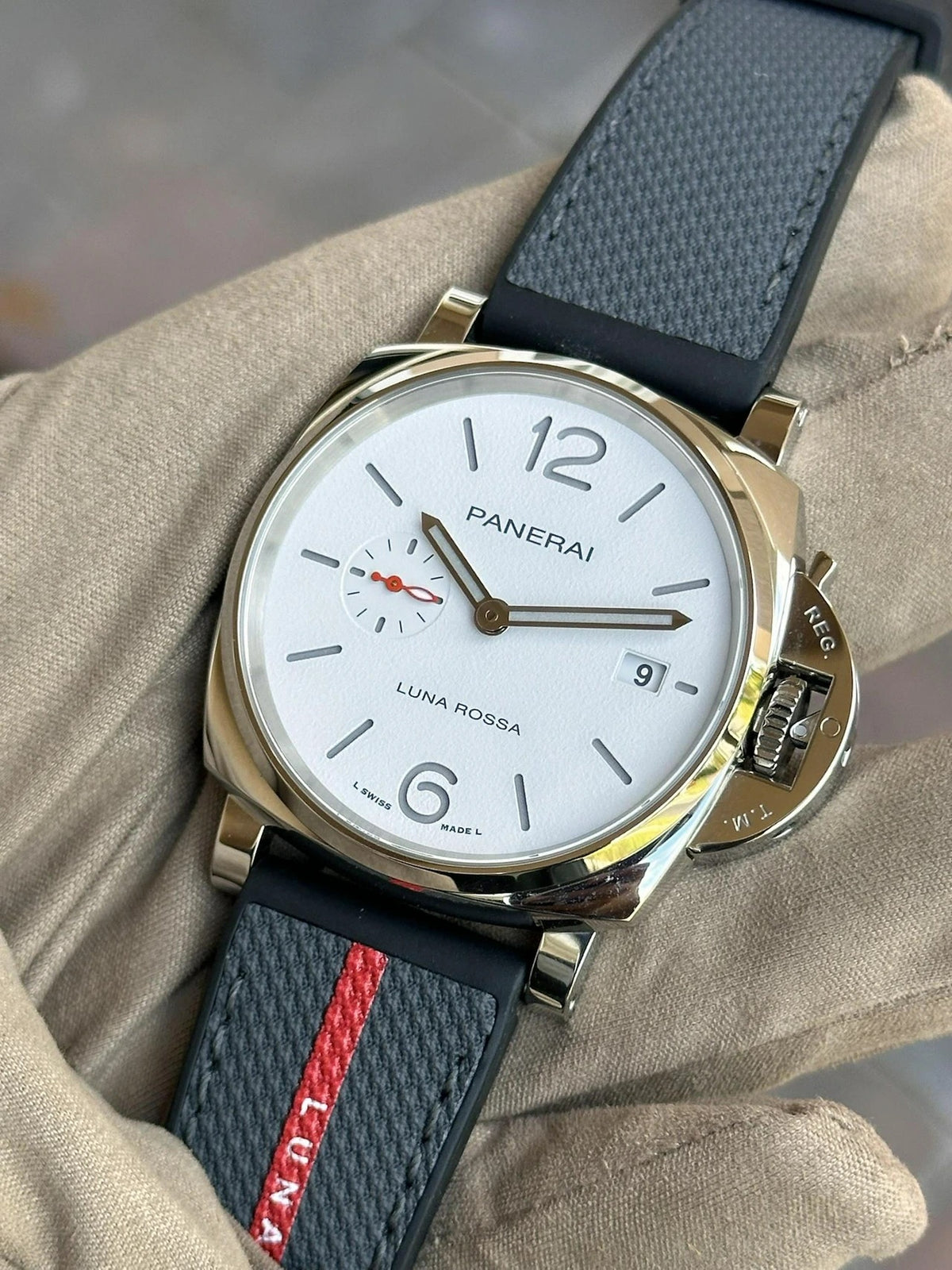 A close-up of a Panerai Luminor Due Luna Rossa PAM01381 wristwatch with a white dial, black leather strap featuring a red stripe, and silver case, held by a hand wearing a beige glove—an ideal timepiece for sailing enthusiasts.