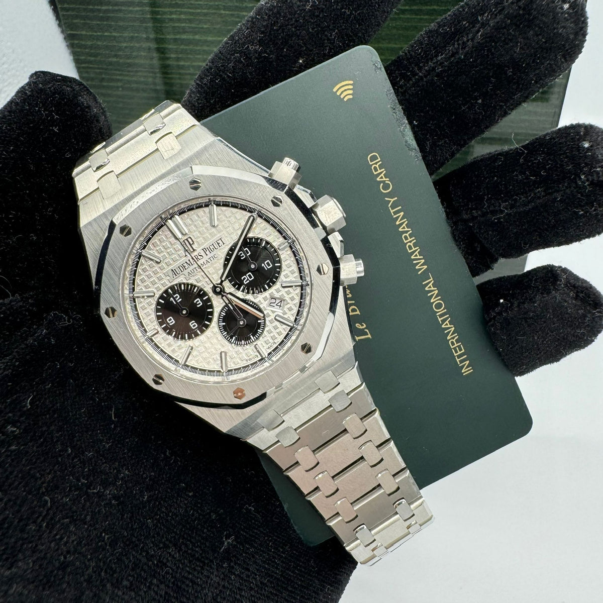 A hand wearing a black glove holds an Audemars Piguet Royal Oak Chronograph 26331ST.OO.1220ST.03, showcasing its stainless steel case, along with an international warranty card.