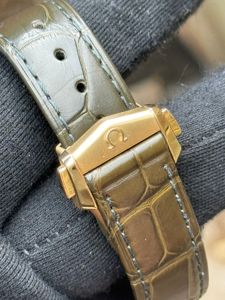 A close-up of a gloved hand holding a watch band with a gold clasp featuring the Omega logo. The strap, brown with a crocodile texture, complements the Omega Speedmaster Professional "Moonshine" 310.63.42.50.10.001 gold case and houses the impressive 3861 manual winding calibre by Omega.