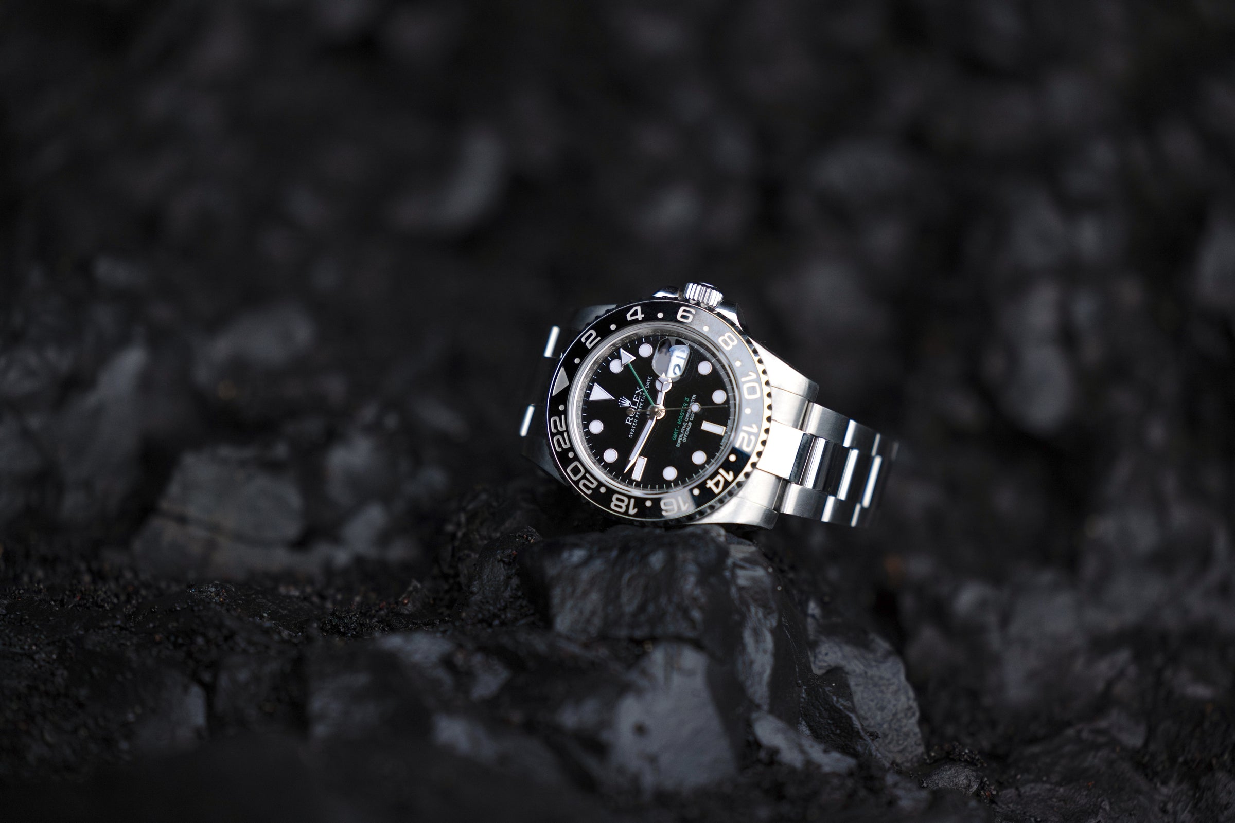 A silver Rolex with the numbers 2 through 22 in increments of two around the bezel. The Rolex has a green hand and is sitting on black volcanic rock.