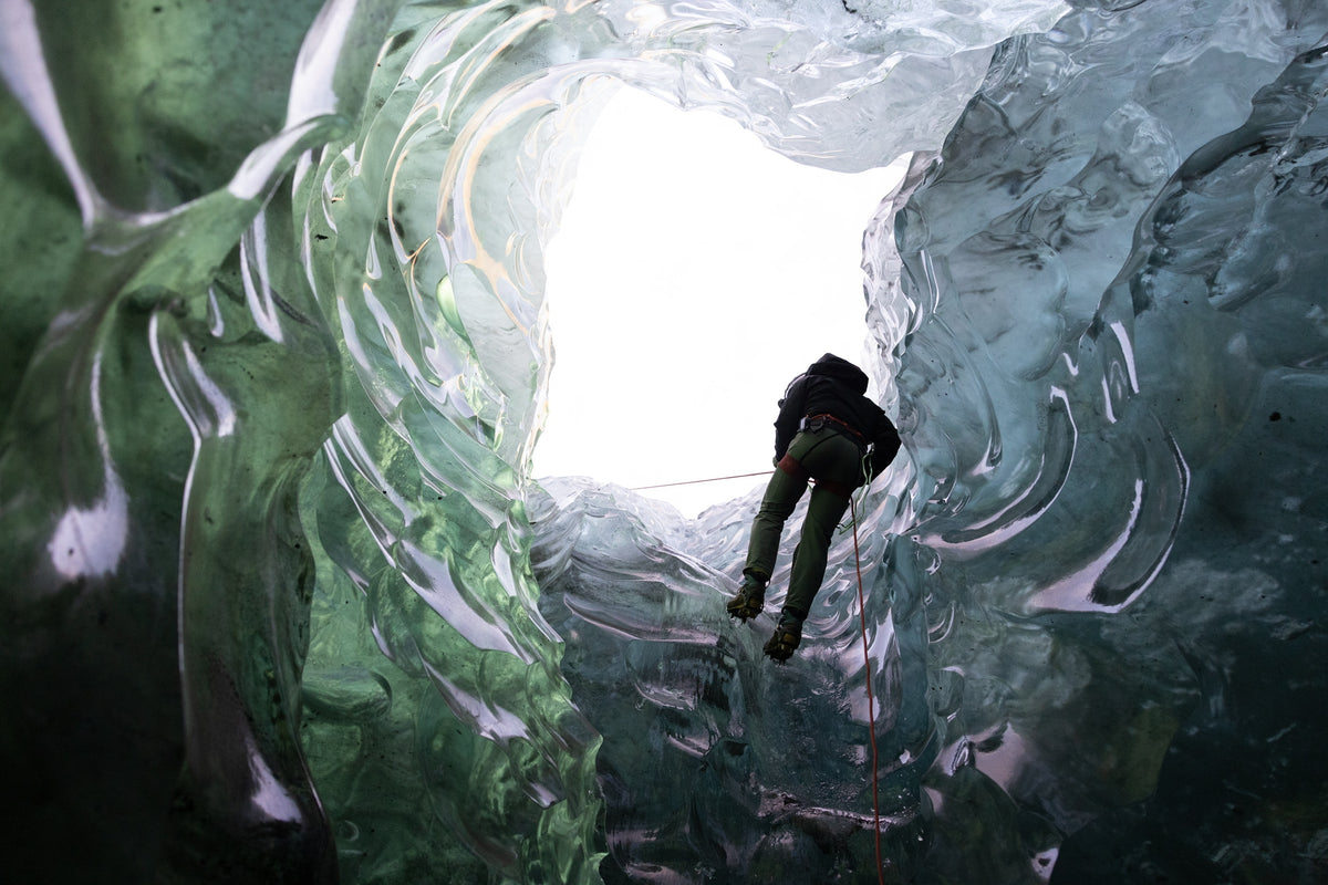 A view from the bottom of an ice cavern of a man climbing glossy green, blue, and clear ice using red rope.