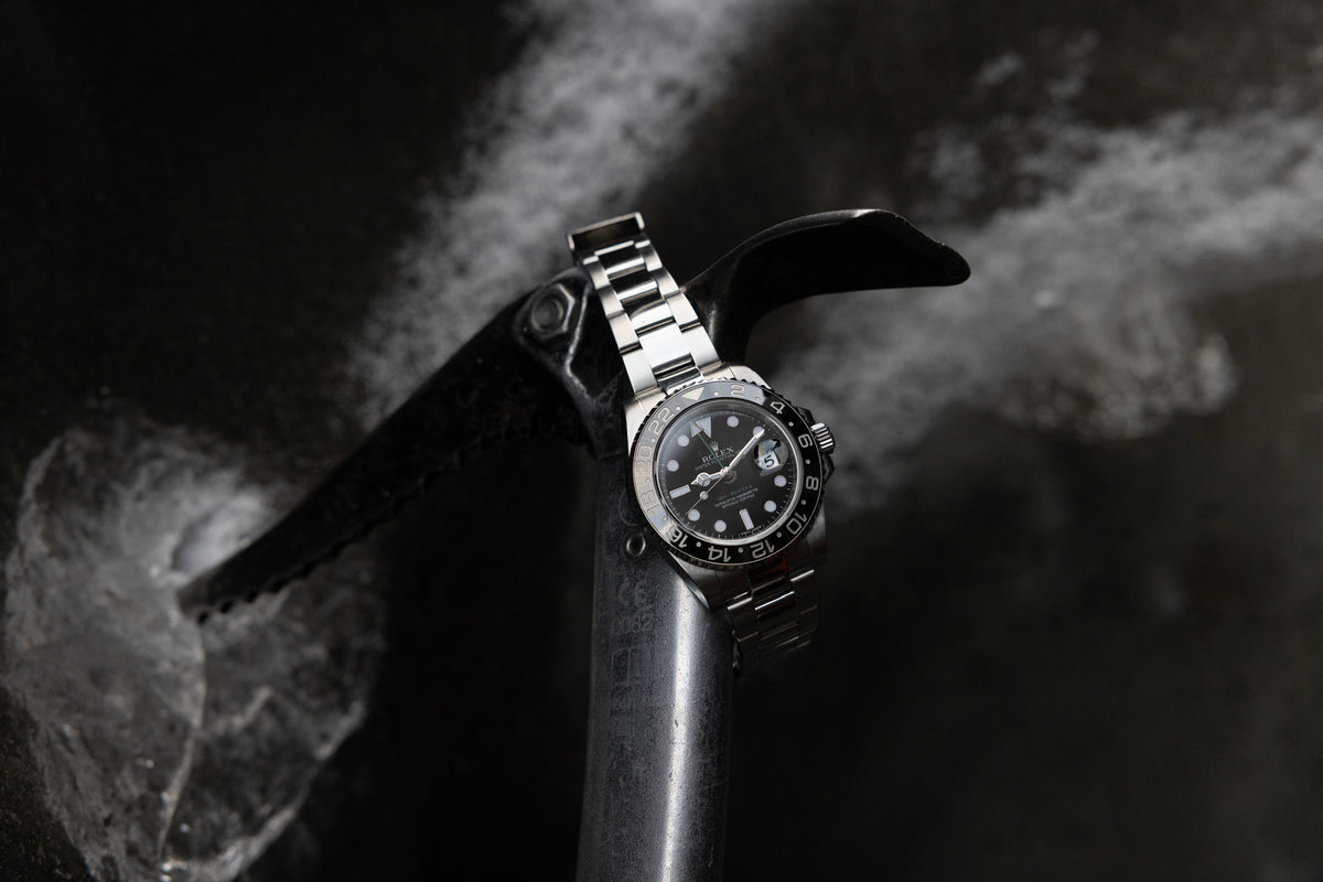 A silver rolex with a black bezel and watch face on a black ice pick that is chiseled into ice.