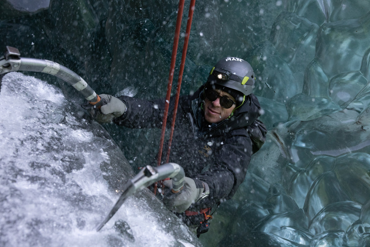 A man climbl ice using two ice hooks while strapped in a harness with red rope, he wears a helmet, sunglasses, a black jacket, and a watch.