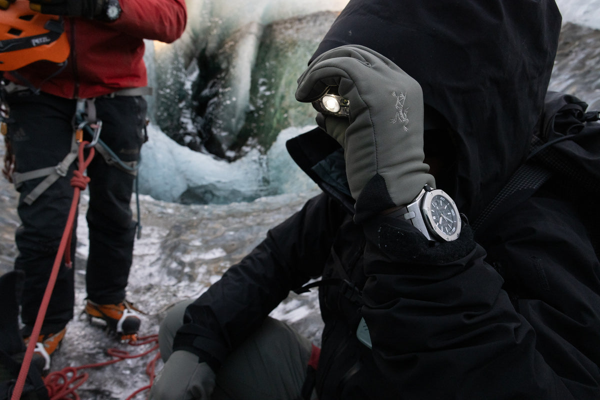 A man wearing a black jacket, gray gloves, and a silver watch outside in ice climbing gear using a headlamp. There is also a person wearing a red jacket holding an orange helmet on the left side of the screen attached to a rope.