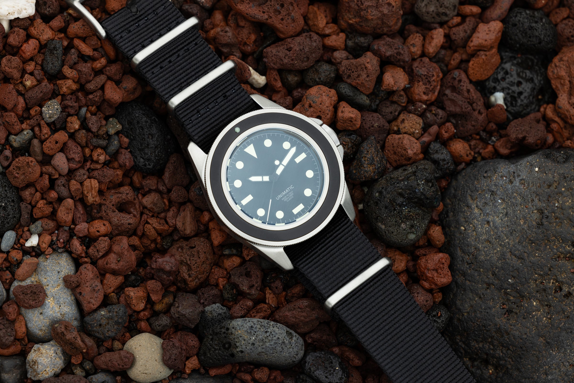 A silver Unimatic watch with a black watch face, bezel, and fabric band lays on brown, black, and gray rocks.